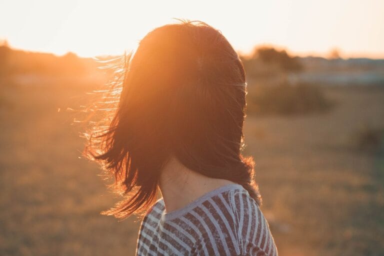 In an open field, a person with shoulder-length hair gazes at the sunset, their striped top gently catching the evening light. They stand strong and resilient, embodying hope akin to those seeking support through domestic violence resources.