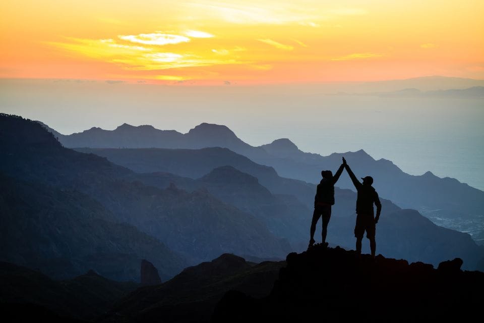 Two people high-fiving on a mountain peak at sunset, with layered mountain ranges and a colorful sky in the background.