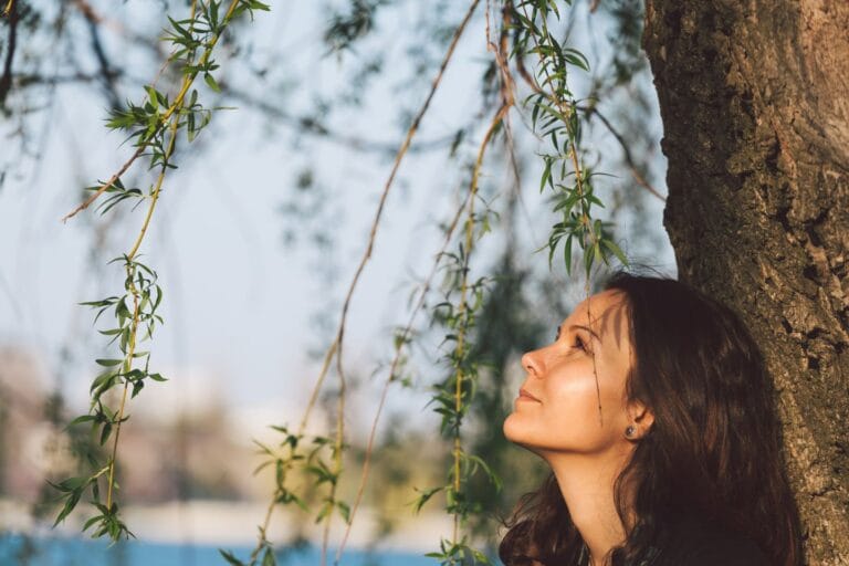 A woman with long hair leans against a tree, looking up at hanging branches in a sunlit outdoor setting.