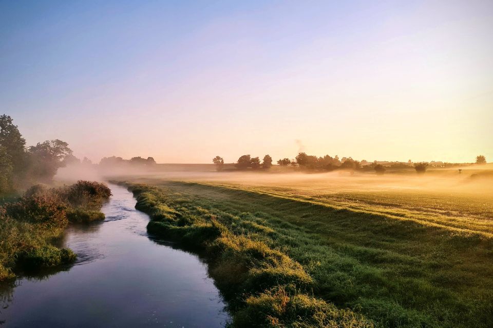 A serene landscape with a calm river flowing through grassy fields and trees under a clear sky at sunrise, with light mist hovering over the ground.