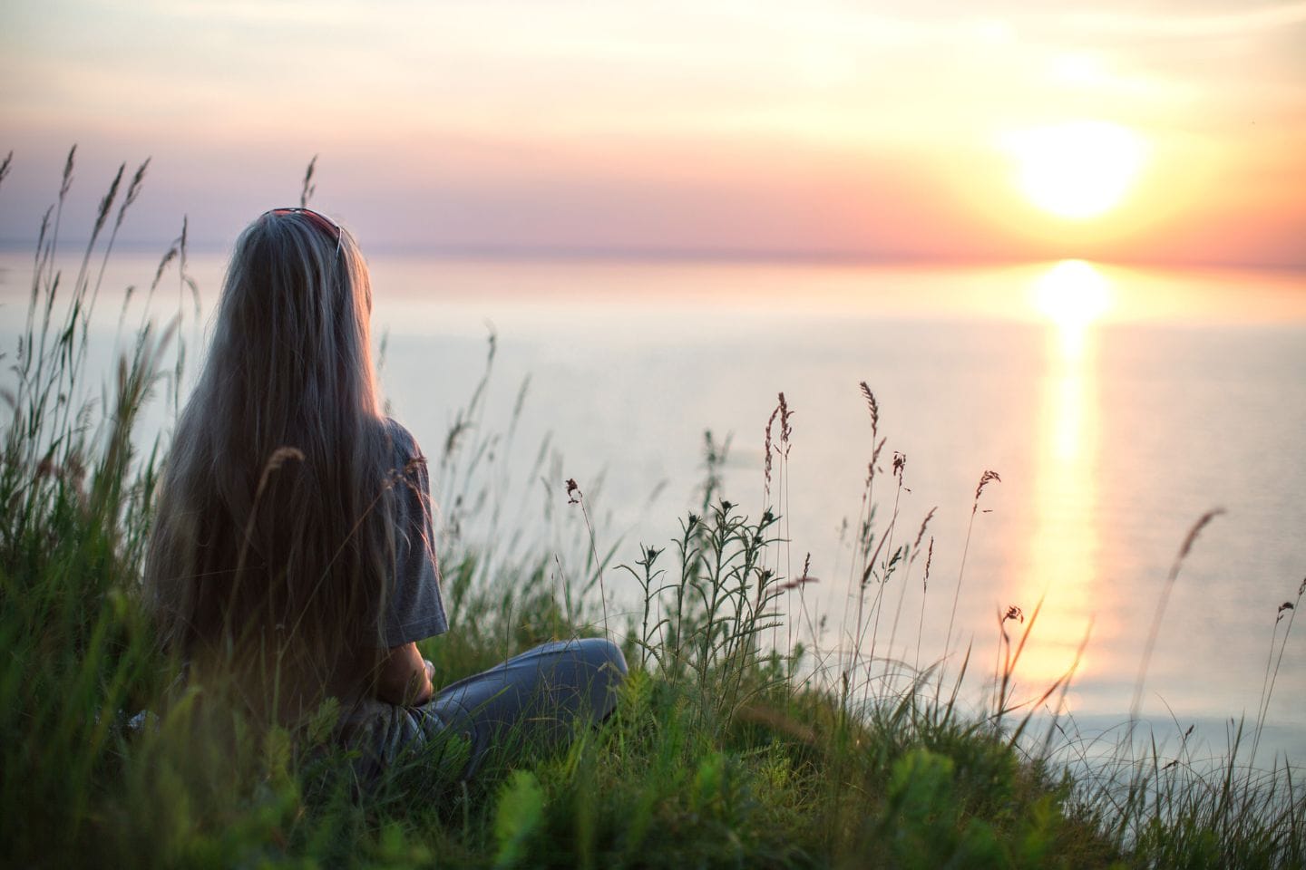A person with long hair sits on grass, facing a calm body of water during a sunset, finding solace and strength from nature's embrace, reminiscent of the resilience built through accessing domestic violence resources.