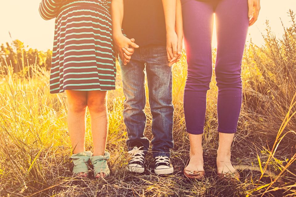 Three people standing in a field, showing only their legs. A child in a striped dress is on the left, a child in jeans and sneakers is in the middle, and an adult in purple pants is on the right.