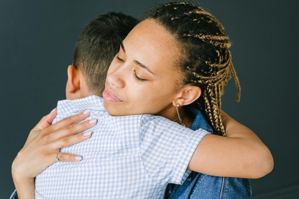 A woman with braided hair hugs a child in a light checkered shirt against a dark background at Jaclyns Place, both facing opposite directions.