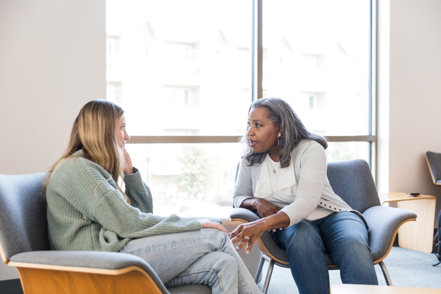 Two women sit in chairs facing each other in a bright room, engaged in a serious conversation. One woman is speaking about available resources while the other listens attentively.