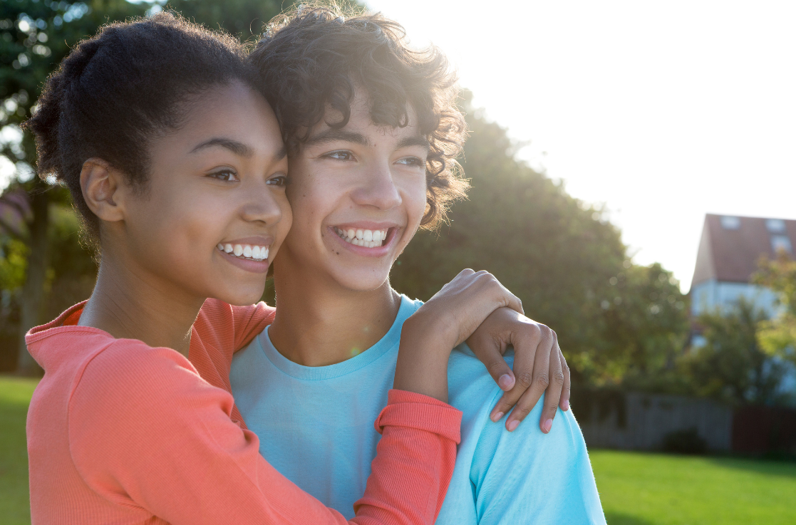 Two teenagers stand together outdoors, smiling and hugging, with trees and a house in the background on a sunny day, enjoying each other's company and the natural resources around them.